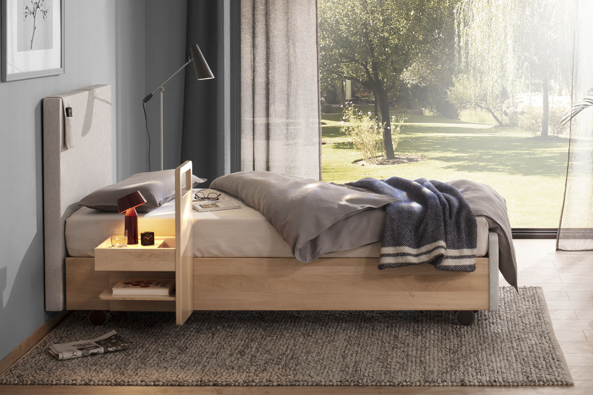 Ein minimalistisches Schlafzimmer mit einem Holzbett, grauer Bettwäsche, blauer Decke, Nachttisch und Stehlampe, durch große Fenster fällt Licht auf einen grünen Garten.