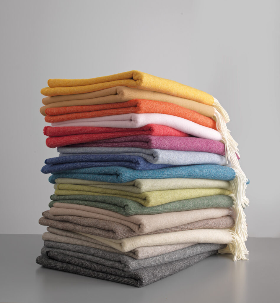 Ein Stapel aus 15 farbigen Decken, von oben nach unten: gelb, orange, rot, rot-pink, rosa, hellblau, türkis, blau, hellgrün, gelbgrün, olivgrün, beige, hellgrau, grau und dunkelgrau, auf einem Tisch gestapelt.