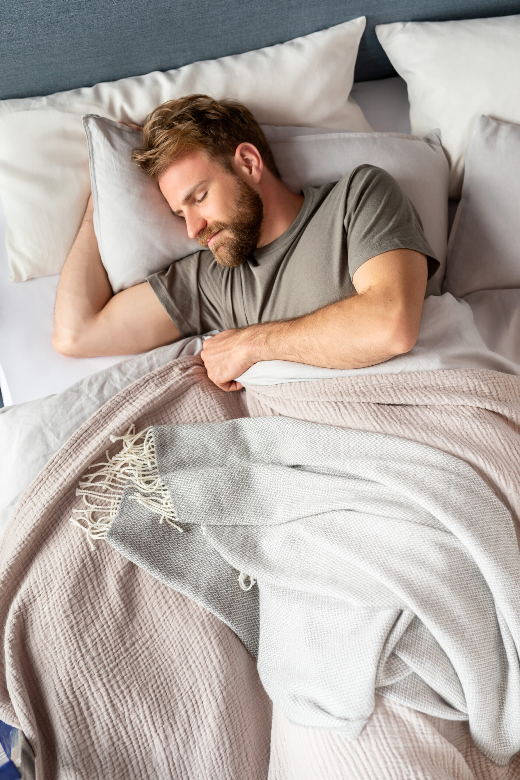 Mann mit Bart schläft friedlich unter einer hellgrauen Decke in einem Bett mit mehreren Kissen.