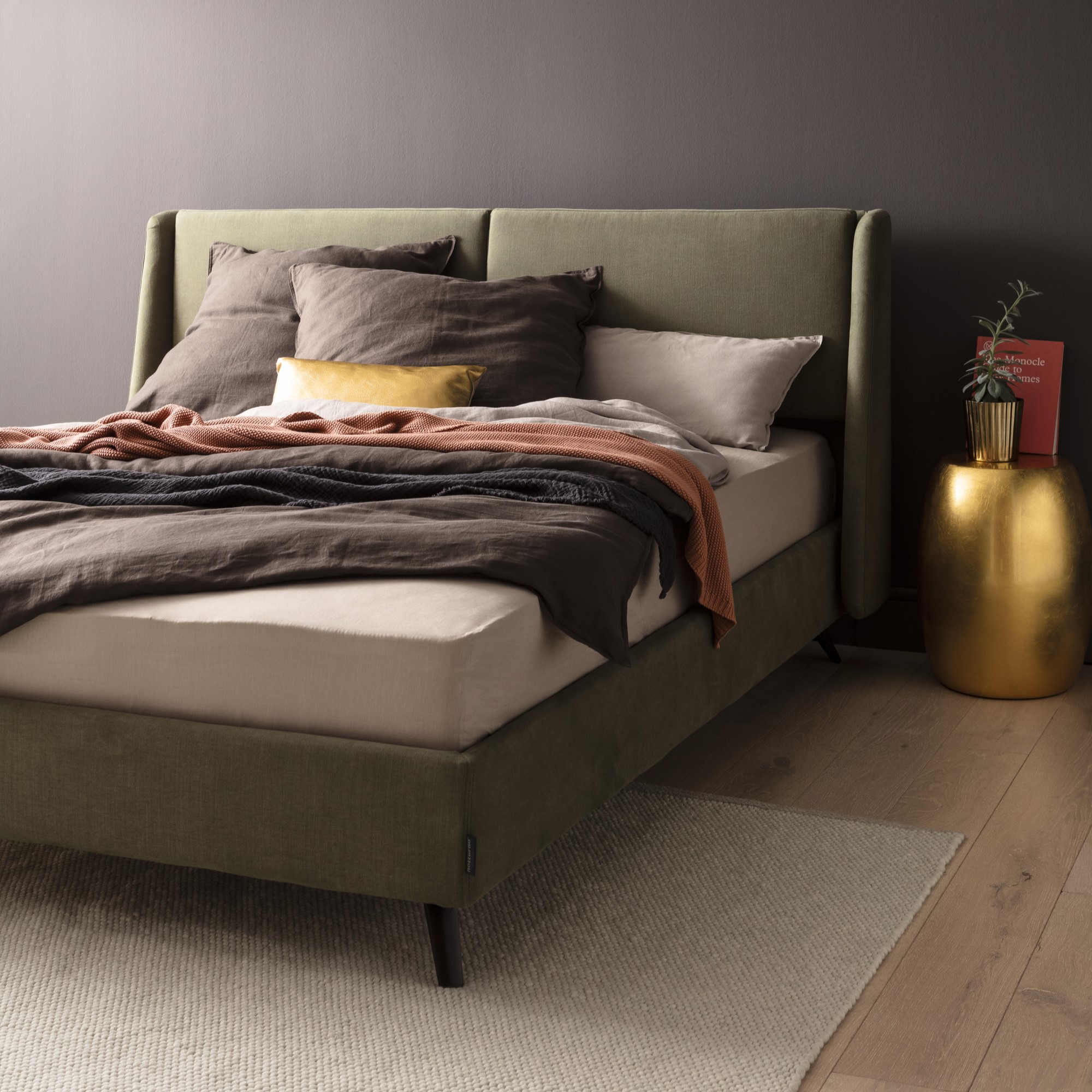 Modernes Schlafzimmer mit olivgrünem Bett, braunen und grauen Kissen, einem roten Buch und einer Pflanze auf einem goldenen Beistelltisch, auf hellem Holzboden mit Teppich.