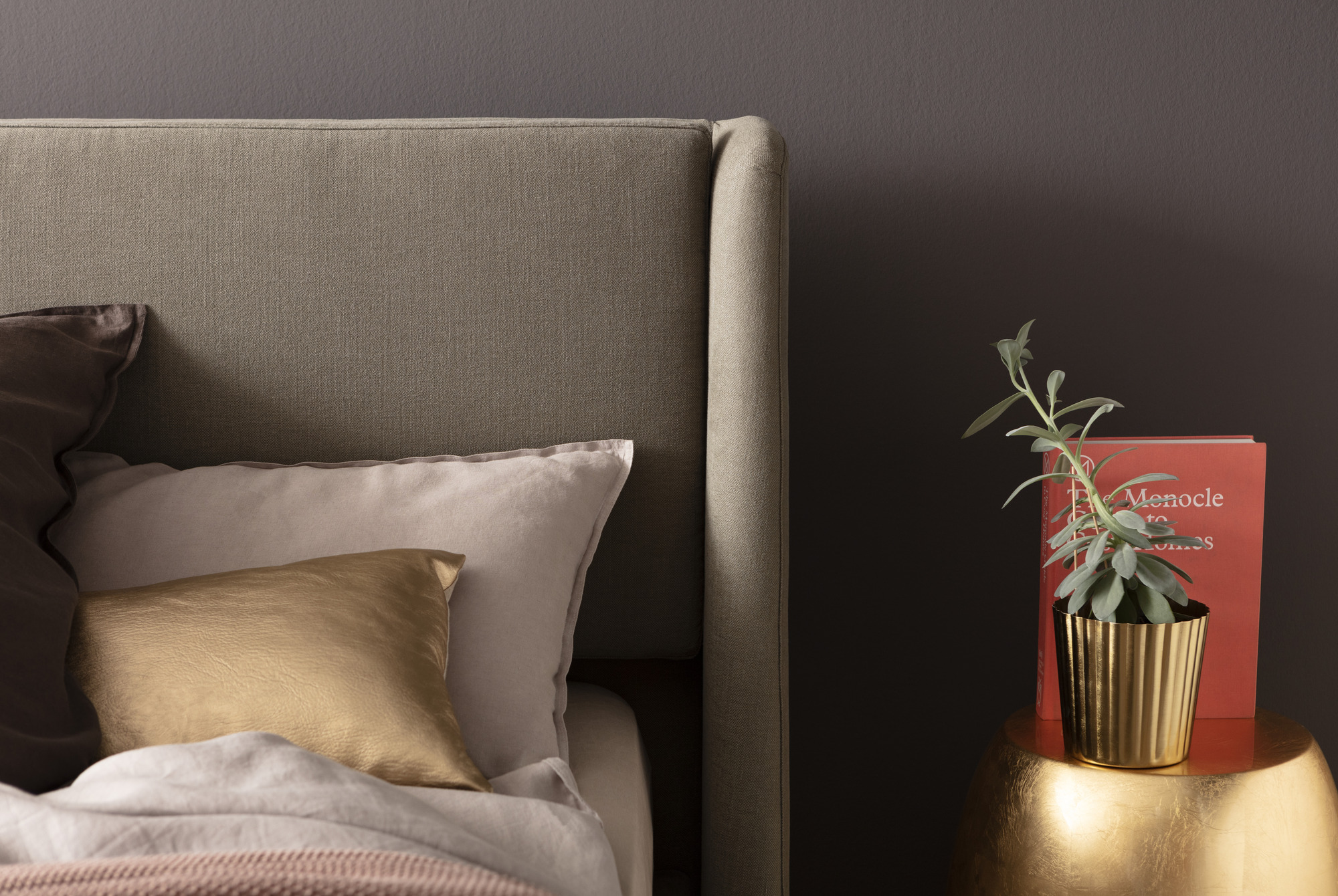 Ein Schlafzimmer mit einem grauen Polsterbett, das mit verschiedenen Kissen in Gold- und Beigetönen dekoriert ist, daneben ein kleiner Tisch mit einer goldfarbenen Pflanzentopf und einem roten Buch.