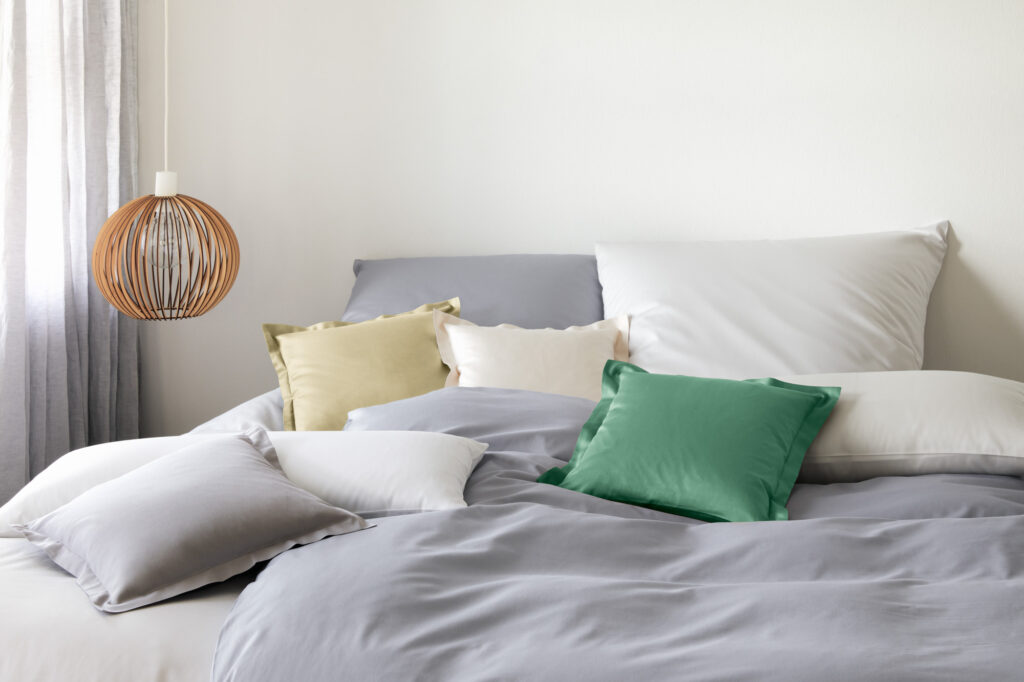 Gemütliches Schlafzimmer mit einem Bett voller Kissen in verschiedenen Farben, darunter grau, grün und beige, neben einer modernen Hängelampe aus Holz und einem halbtransparenten Vorhang.