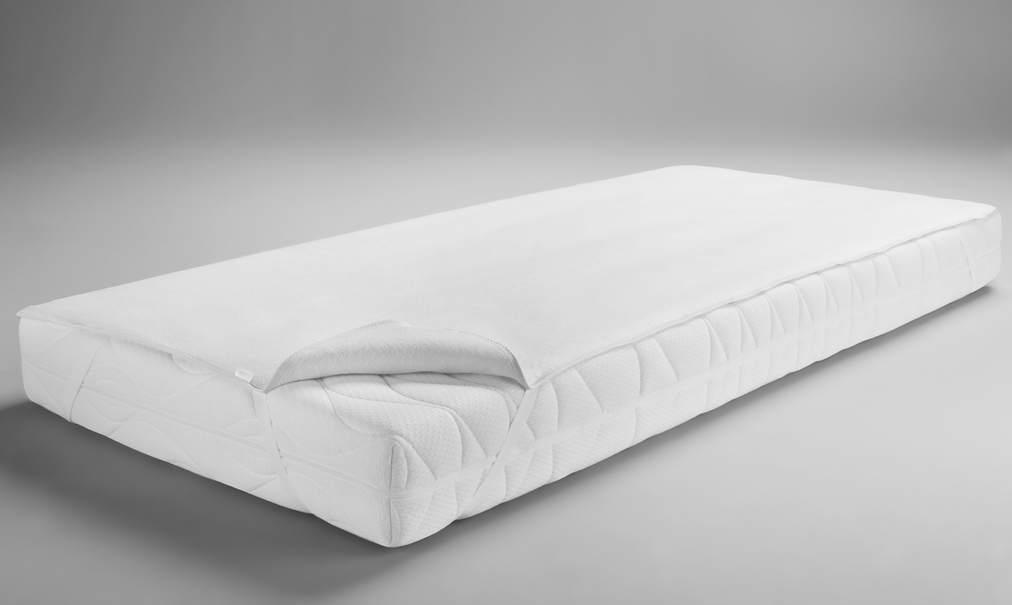 Weißer Matratzenaufleger auf einer Matratze mit gestepptem Bezug in einem grauen Hintergrund.