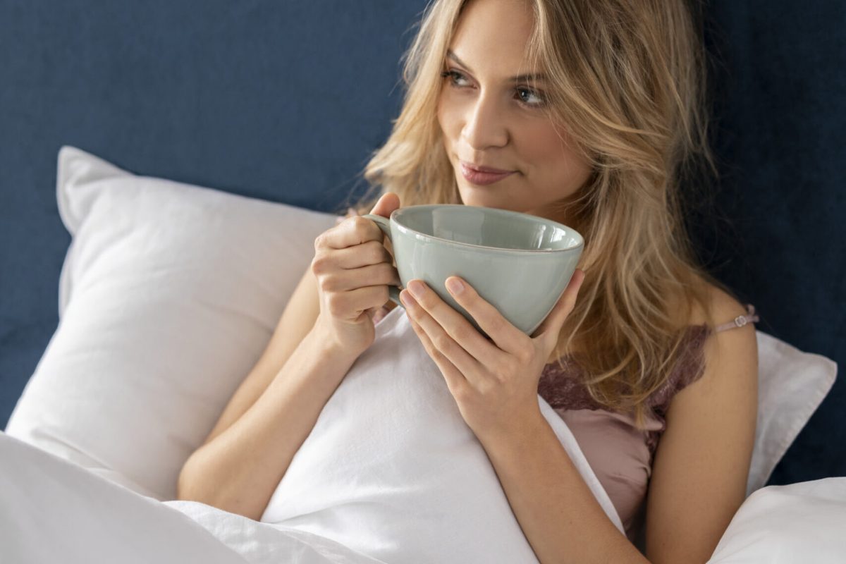 Frau mit blonden Haaren sitzt im Bett, hält eine große Tasse, umhüllt von einer weißen Decke, vor einem blauen Kopfteil.