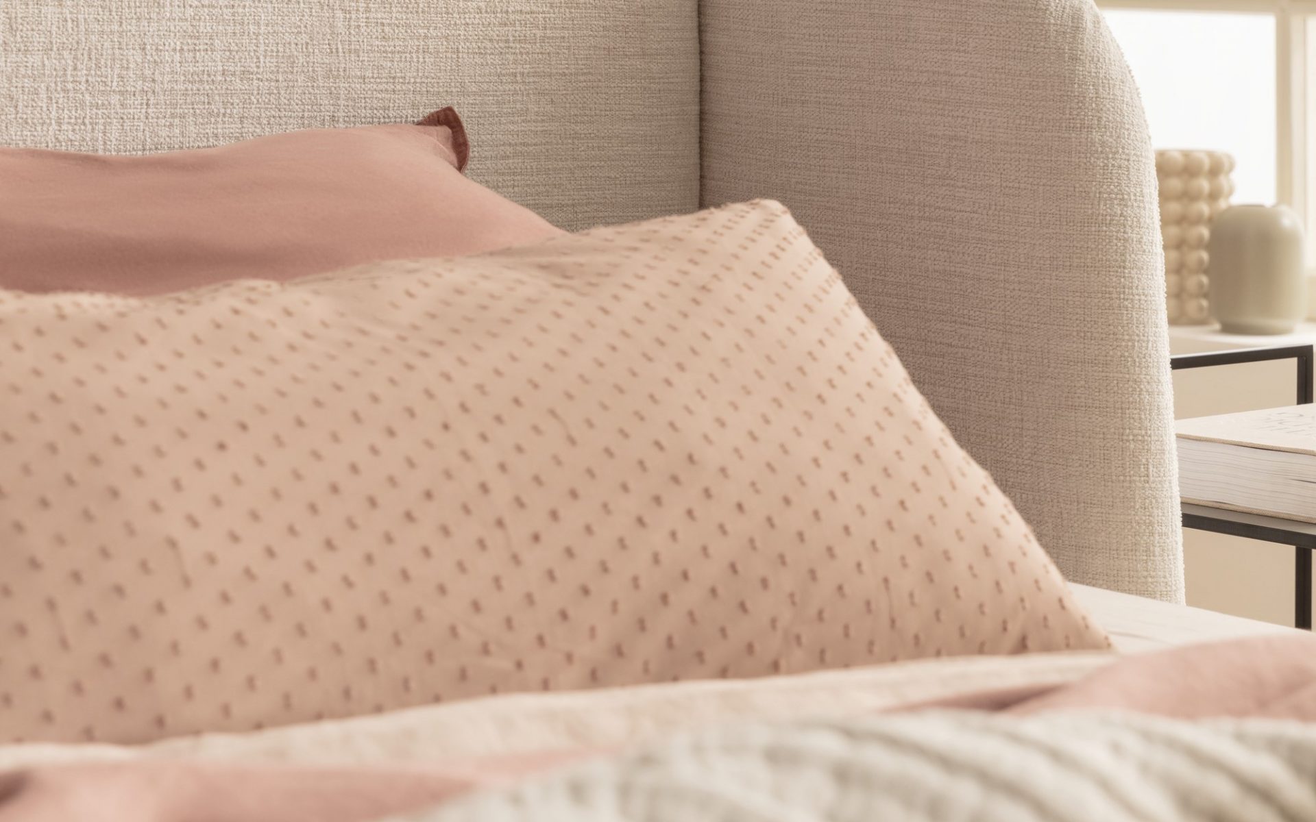Ein Bett mit einem hellen, gepolsterten Kopfteil, weichen rosa und beigefarbenen Kissen und einer gemusterten Decke in einem hellen, gemütlichen Raum.