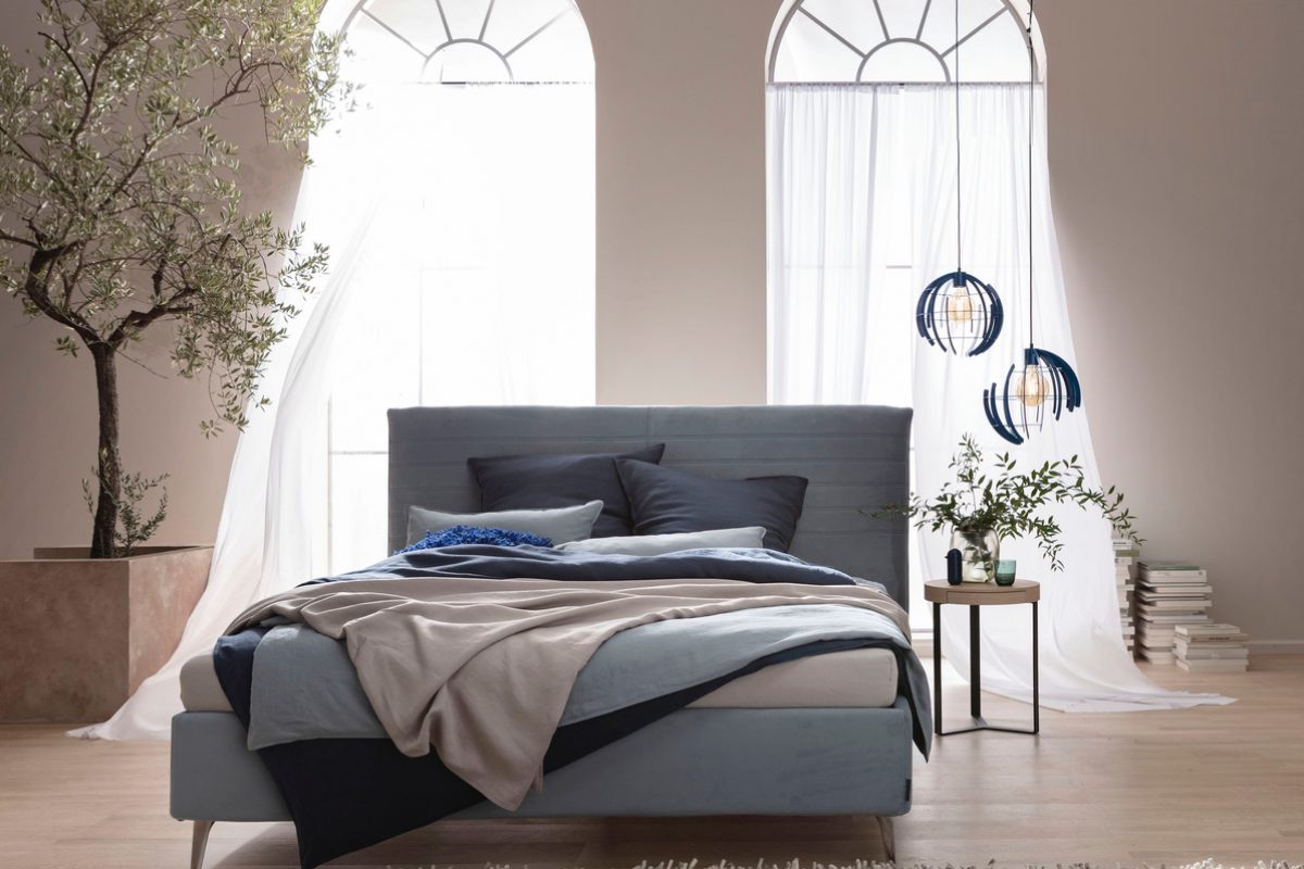 Ein Schlafzimmer mit einem Bett in Grau- und Beigetönen, umgeben von einem Olivenbaum und moderner Beleuchtung. Im Hintergrund sind zwei bogenförmige Fenster mit weißem durchscheinenden Vorhang zu sehen und ein kleiner Tisch mit Pflanzen und Büchern.