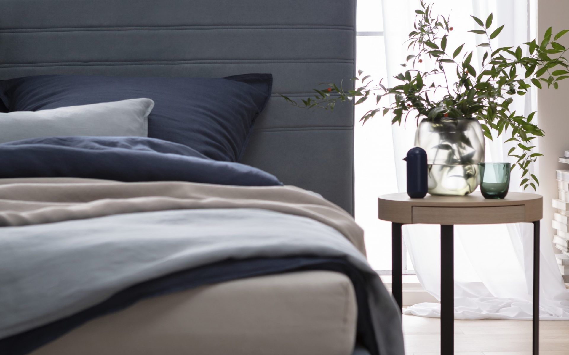 Schlafzimmer mit einem grauen gepolsterten Kopfteil eines Bettes, dunkelblauen und beigen Bettwäsche, und einem kleinen Holztisch mit einer Vase voller grüner Zweige daneben.