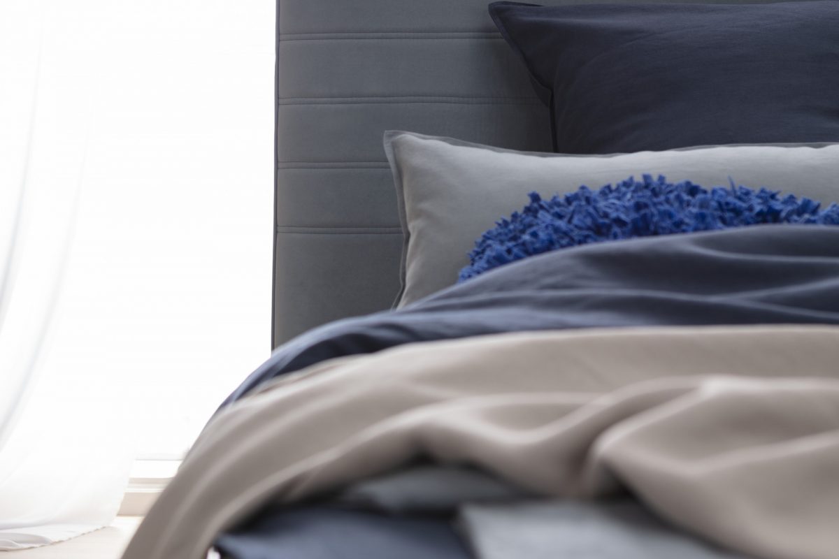 Naheaufnahme eines Bettes mit einem gepolsterten grauen Kopfteil, blauen und grauen Kissen, blauer und grauer Bettwäsche sowie einer beigen Decke.
