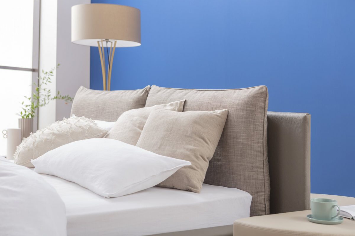 Ein Bett mit hellen Kissen vor einer blauen Wand; daneben steht eine moderne Stehlampe und ein kleines Tischchen mit einer Tasse darauf.